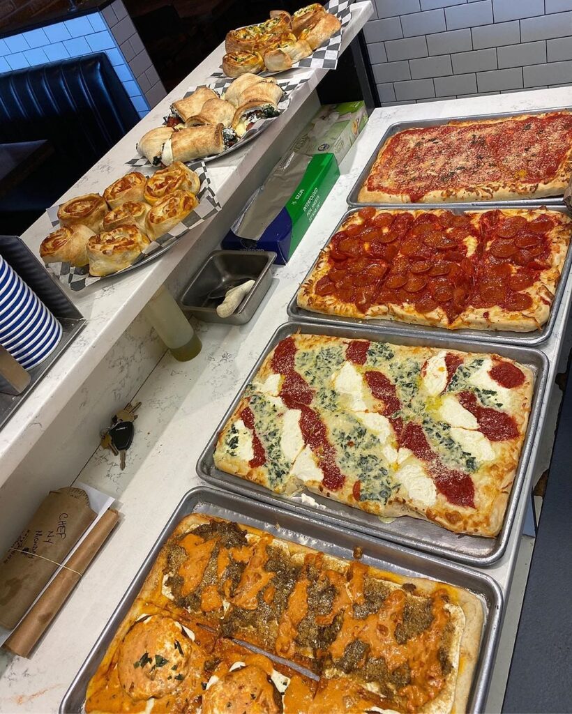 The counter lined with trays of Sicilian pies beside baskets of pinwheels and stuffed rolls
