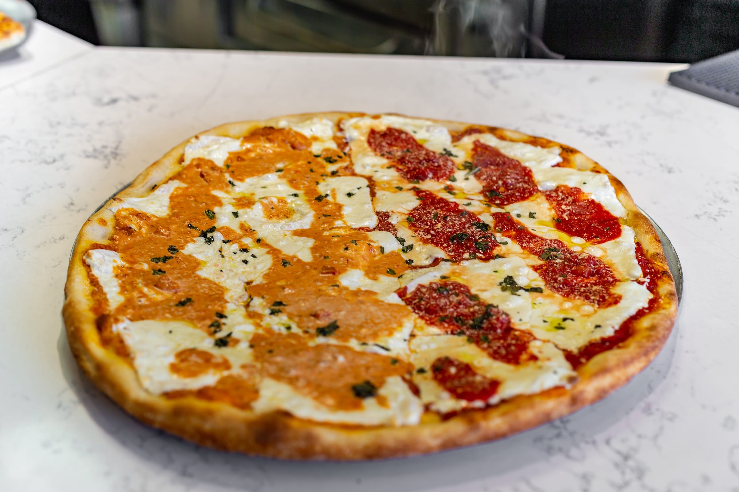 A round pie fresh from the oven, half topped with creamy vodka sauce and half with tomato sauce, both with fresh mozzarella and chopped basil