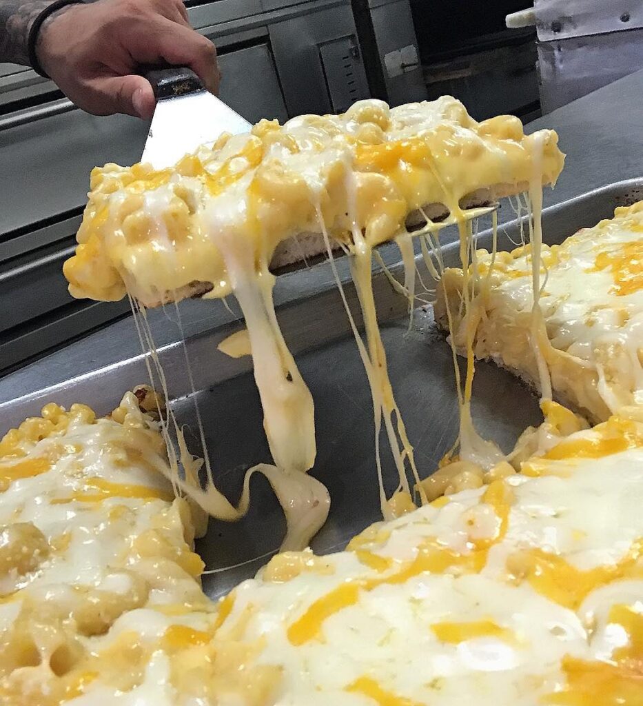 A square of mac and cheese pizza lifted from the tray, with long strands of melted cheese