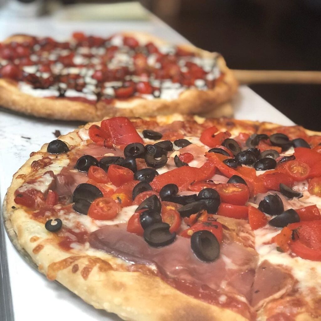 Two individual pizzettas on the counter, one topped with roasted peppers, cherry tomatoes and black olives