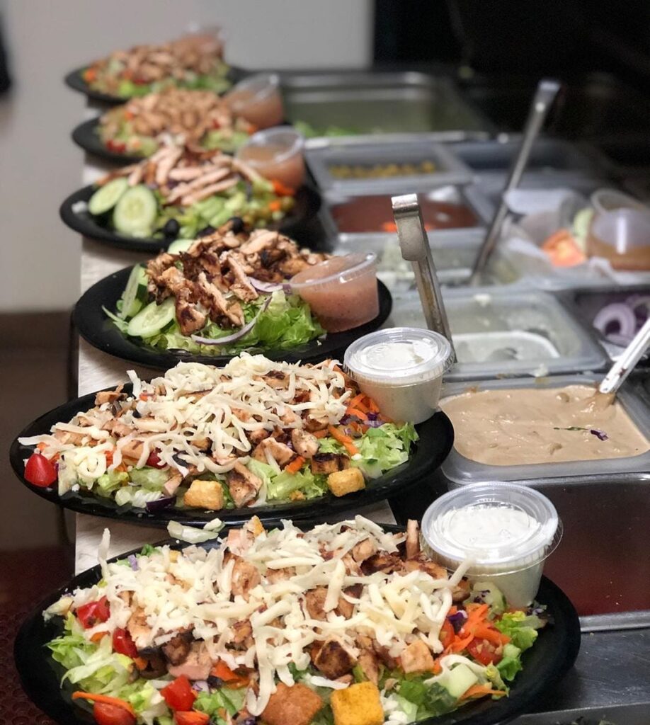 A row of salads topped with grilled chicken and shredded cheese, each with a cup of dressing